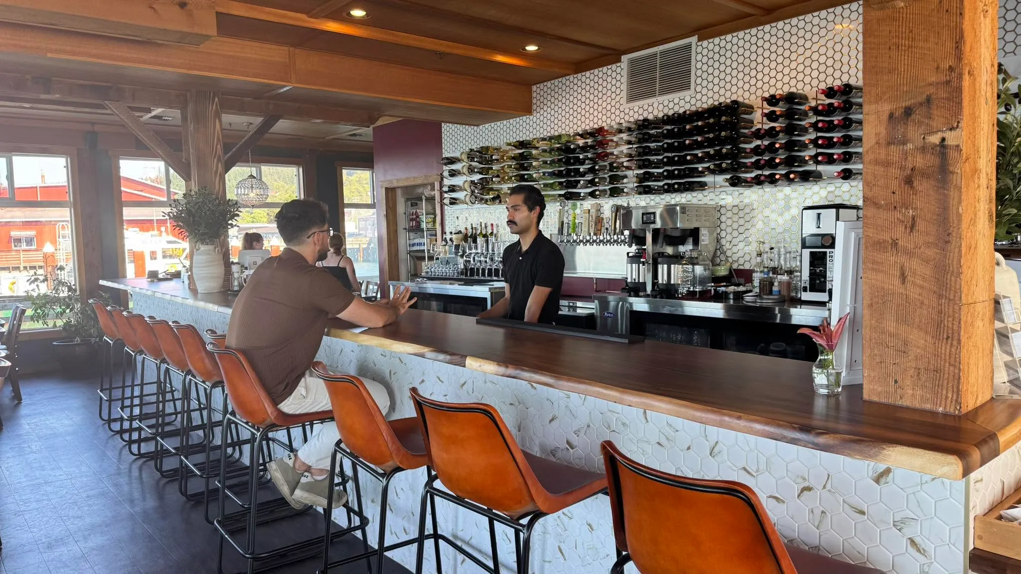 The bar at Akres: leather stools along a white tiled counter, a bartender talking with a guest, wine racked on the wall behind
