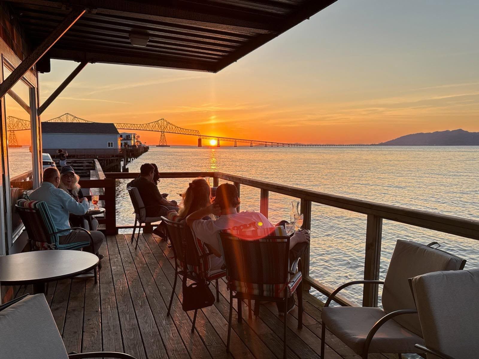 Guests at tables on the covered deck at Akres as the sun sets behind the Astoria–Megler Bridge over the Columbia River