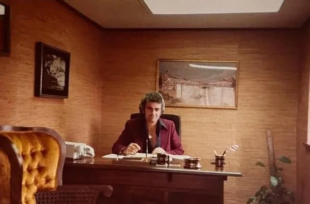 Konstantinos Macheras in a burgundy jacket at a wooden desk in a wood-panelled office