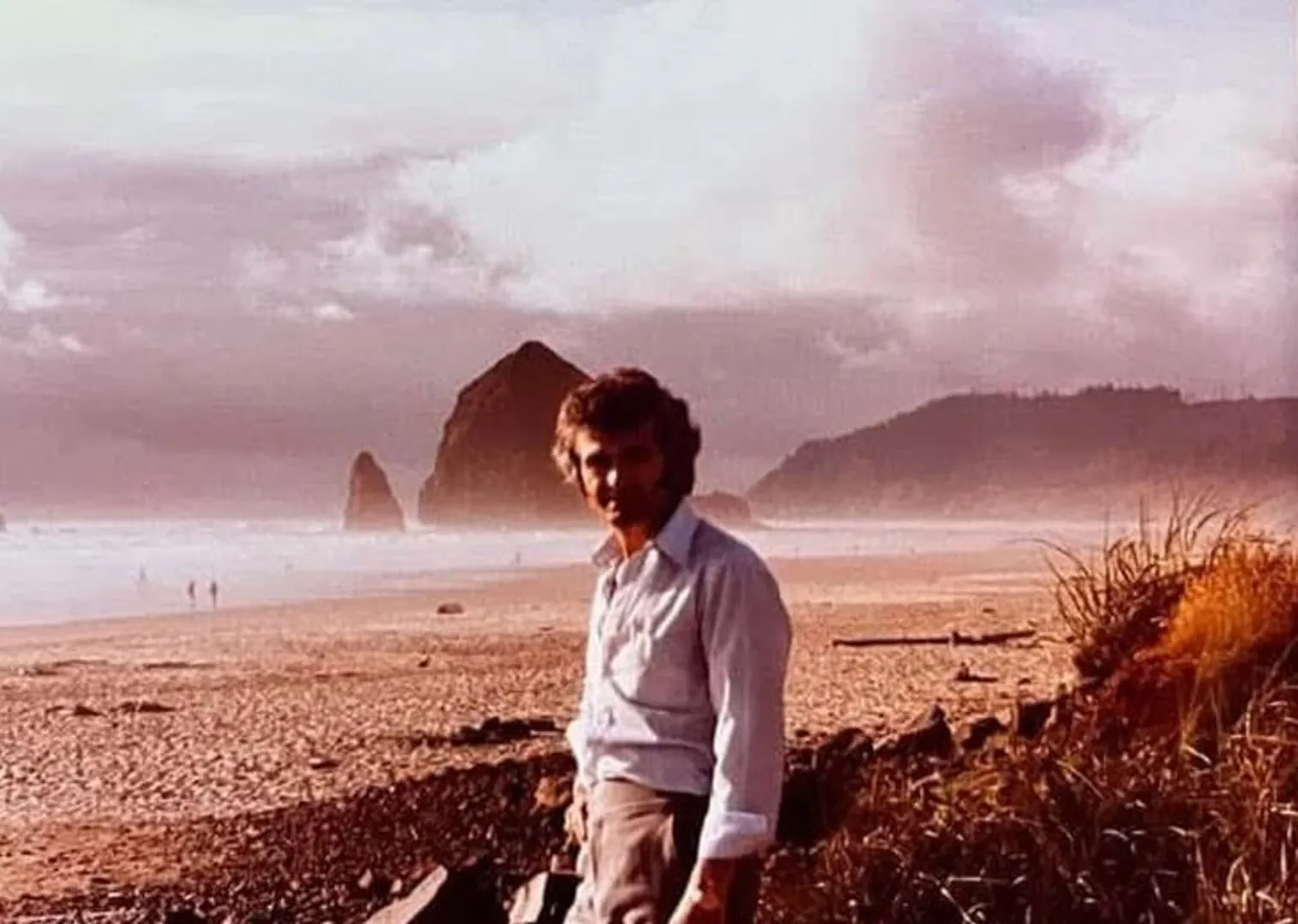Konstantinos Macheras on the beach at Haystack Rock, Cannon Beach, in the 1970s: white shirt, hair in the wind, the rock and the surf behind him